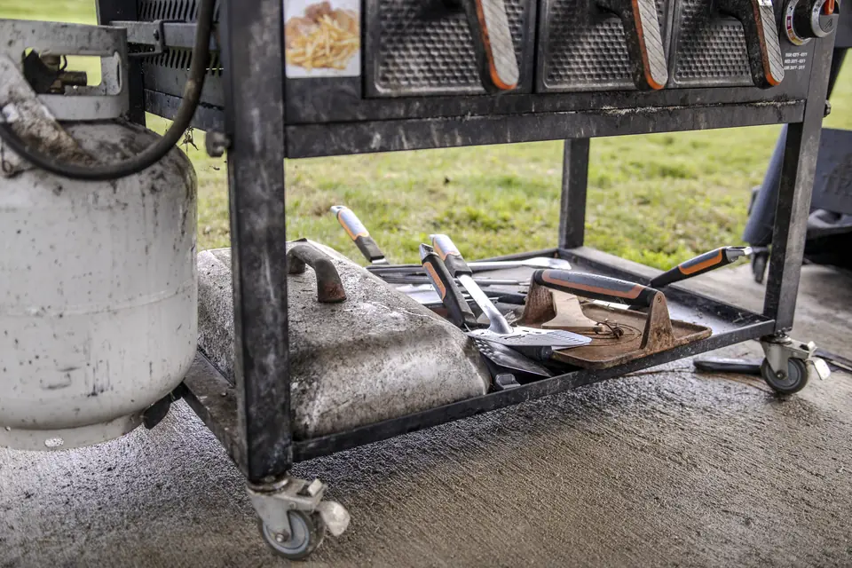 The cluttered bottom shelf of a worn standalone grill cart, with a dirty propane tank and tools piled in